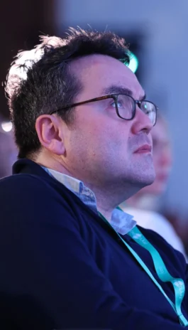 A man with glasses and a lanyard looks attentive during an event.