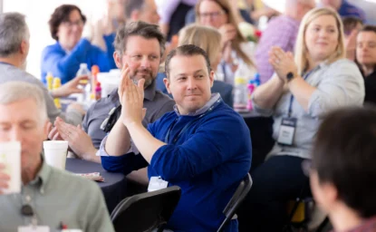 People seated at a gathering, some clapping and others engaged in conversation. Most individuals are wearing name tags. The setting appears to be informal with food and drinks on the tables.