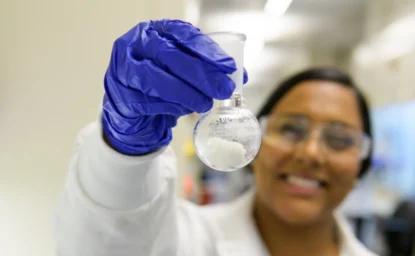 A scientist in a lab coat and safety glasses holds up a flask containing a white substance, smiling and wearing blue gloves.