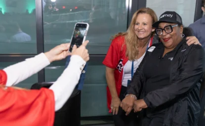 Two women pose for a photo, one wearing a red shirt and the other in a black outfit and glasses. An unseen person takes their picture with a smartphone.